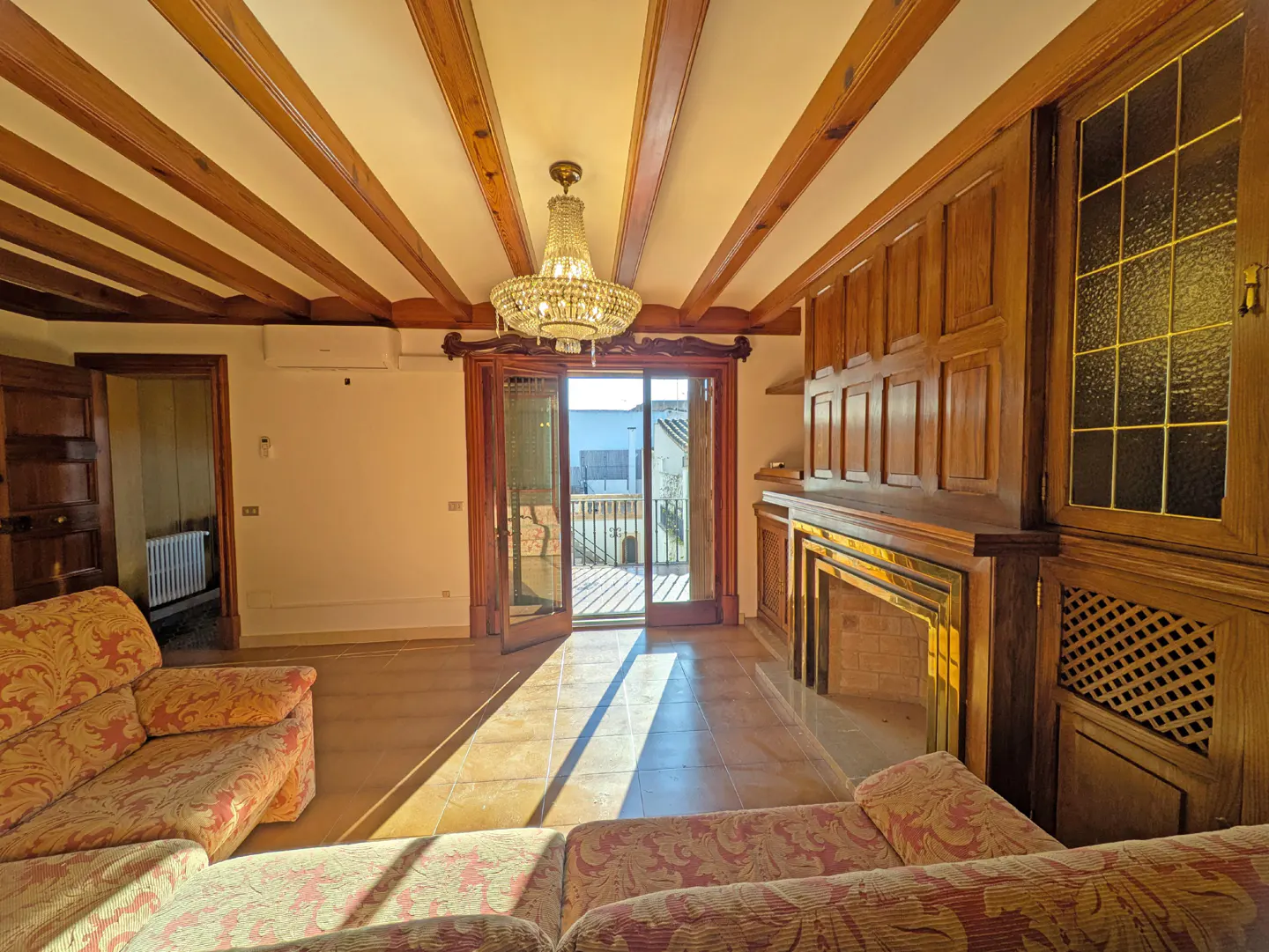 Living room with wood-beamed ceiling, chandelier, fireplace, and patterned sofas. Balcony doors open to a sunny view.