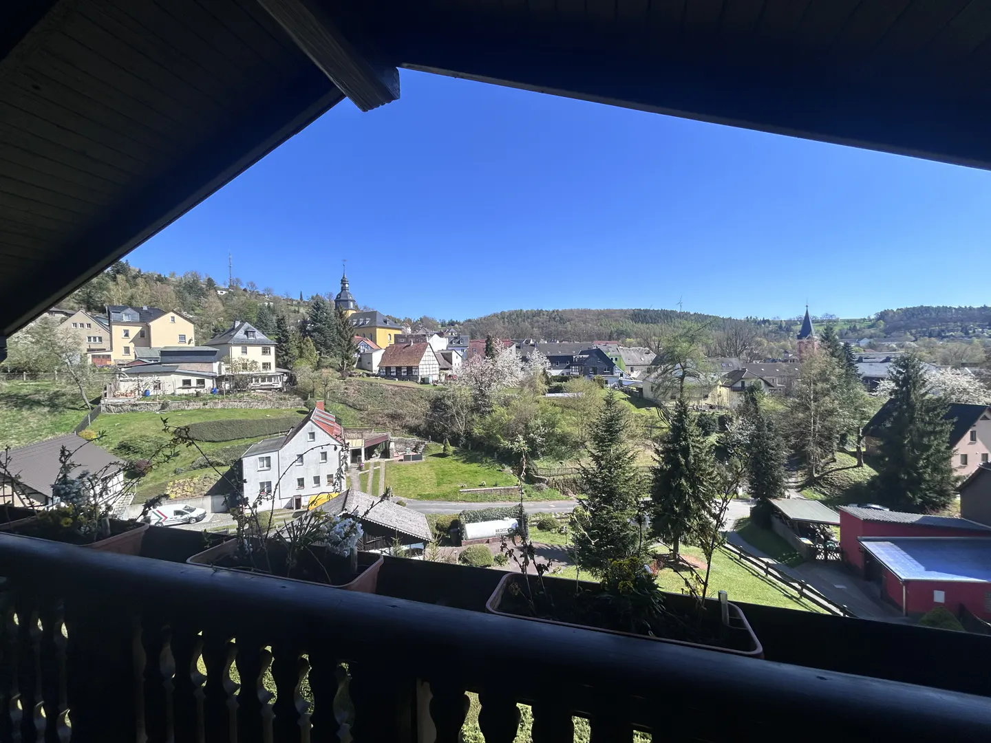 View from a dark wood balcony overlooking a European village on a sunny day. Buildings and trees dot the green hills.