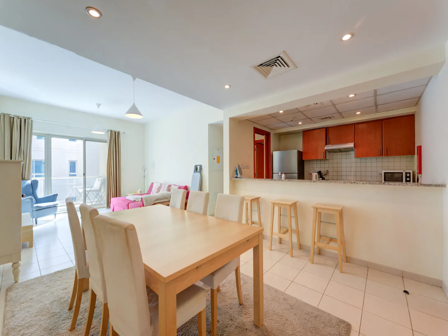 Open-concept living space with a dining table, chairs, and kitchen with bar stools. Balcony with chairs is visible.