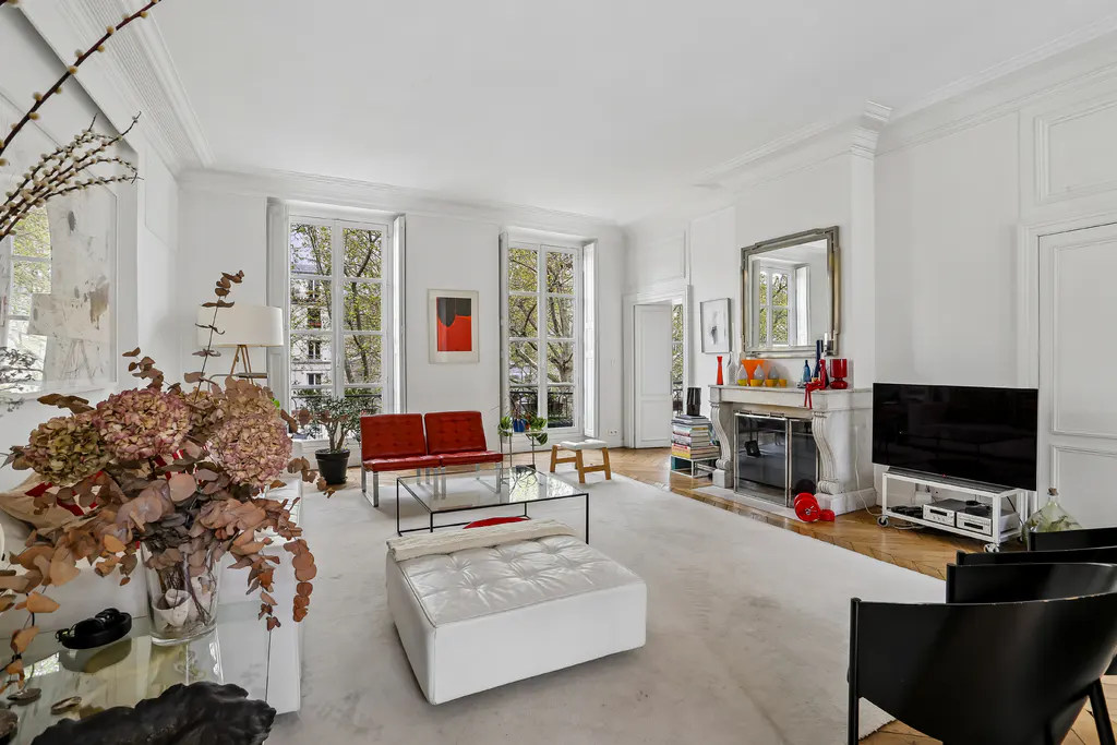 Bright living room with white walls, large windows, and a fireplace. Red sofa, white ottoman, and black chairs furnish the space.