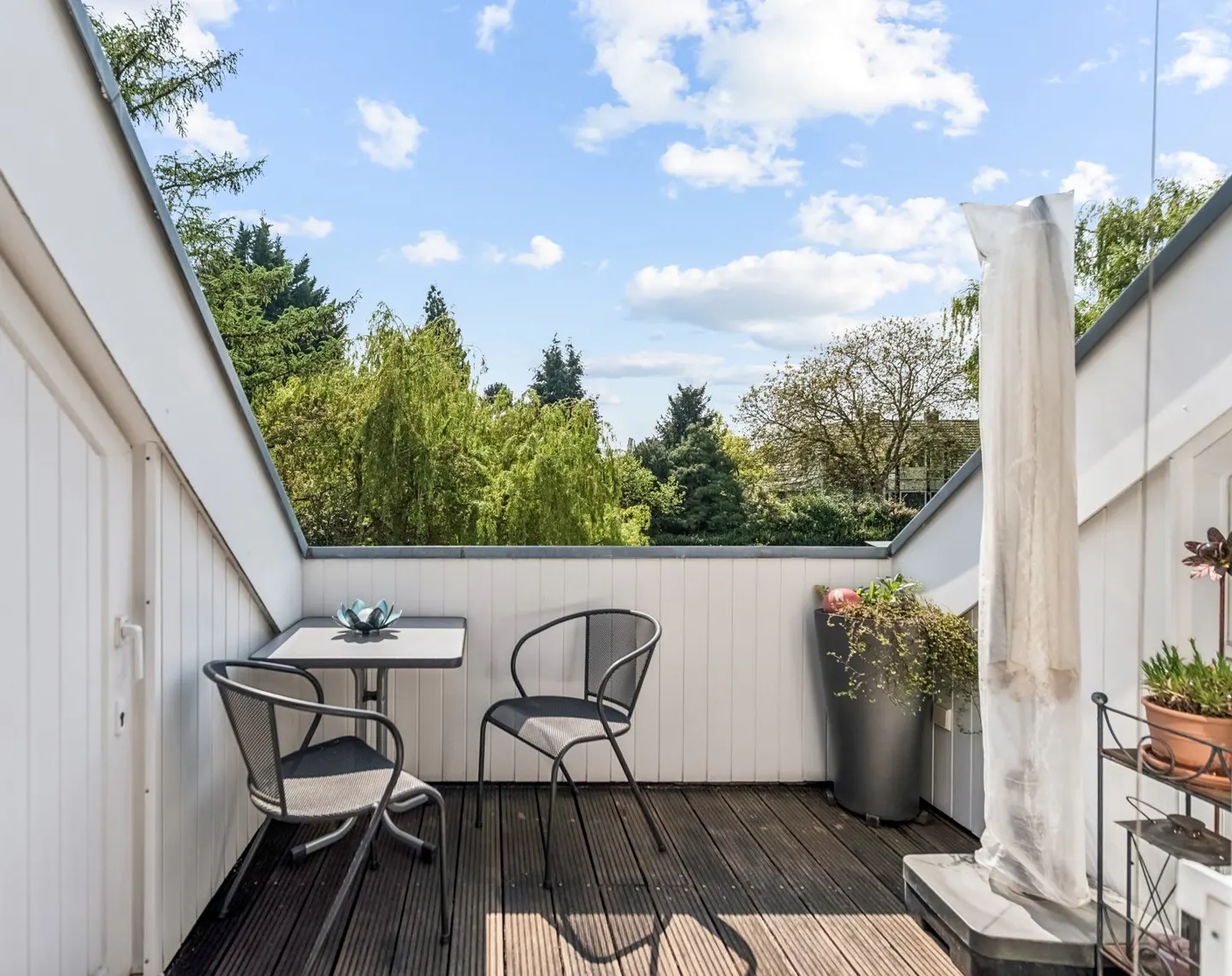 A balcony with a table, two chairs, and potted plants overlooks green trees under a blue sky.