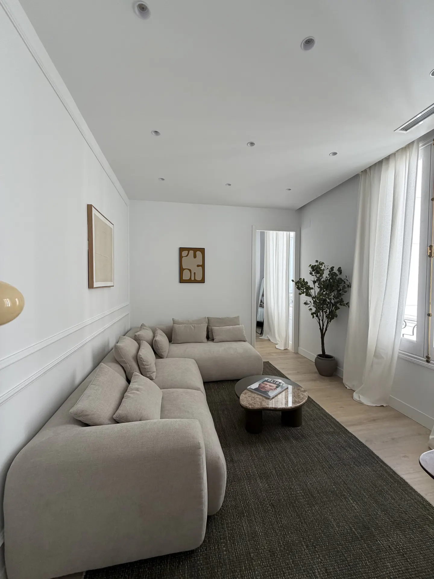 Bright living room with a large beige sectional sofa, a dark rug, and a marble coffee table. White walls, art, and a potted tree add to the minimalist aesthetic.