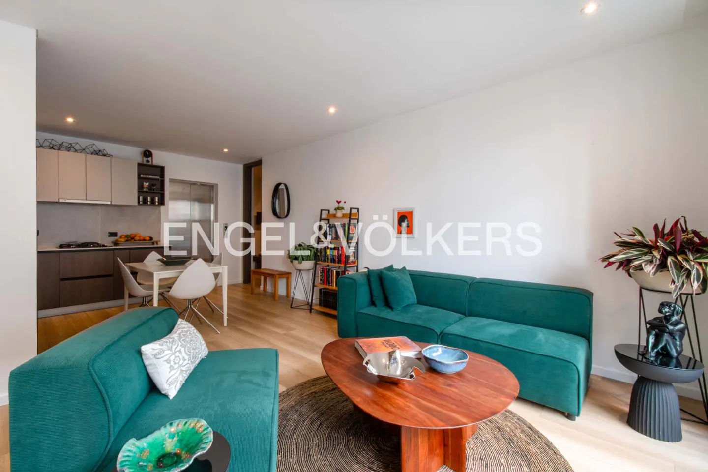 Bright living room with teal sofas, a wood coffee table on a round rug, and a kitchen in the background.
