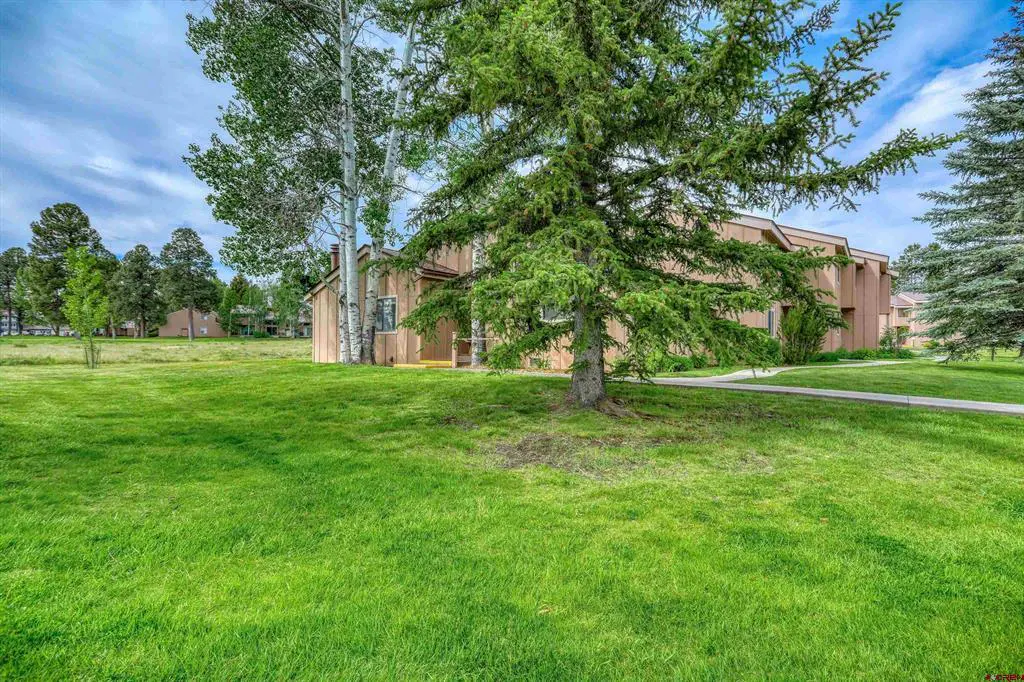 Tan multi-unit residential building surrounded by a large green lawn, tall pine trees, and a blue sky with light clouds.