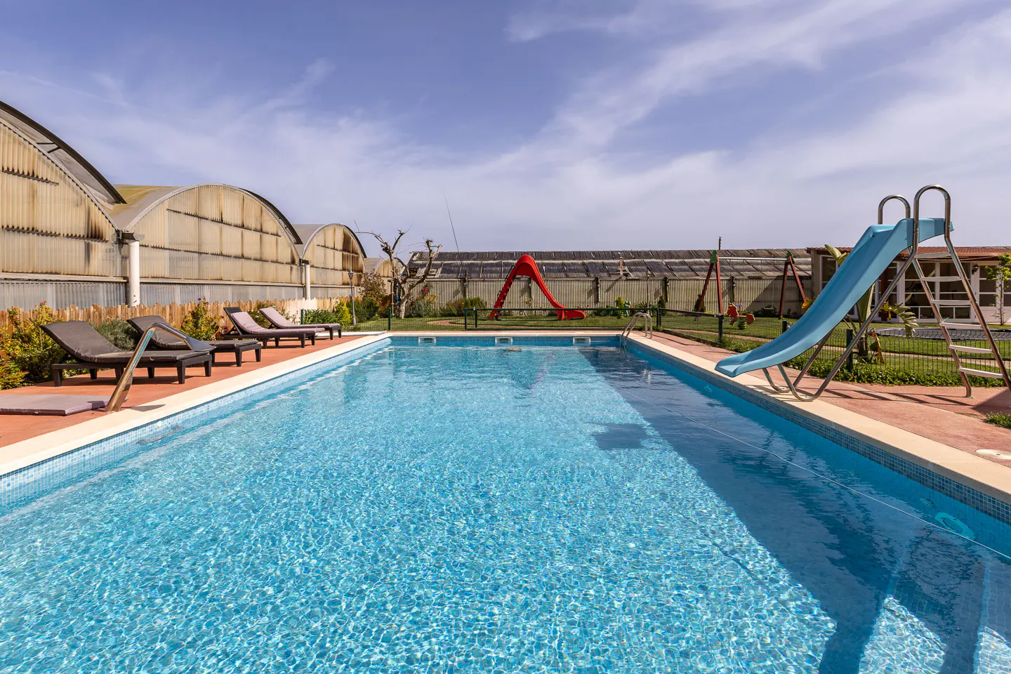 Outdoor pool with blue water, lounge chairs, and a blue slide. A red slide and swingset are in the background.