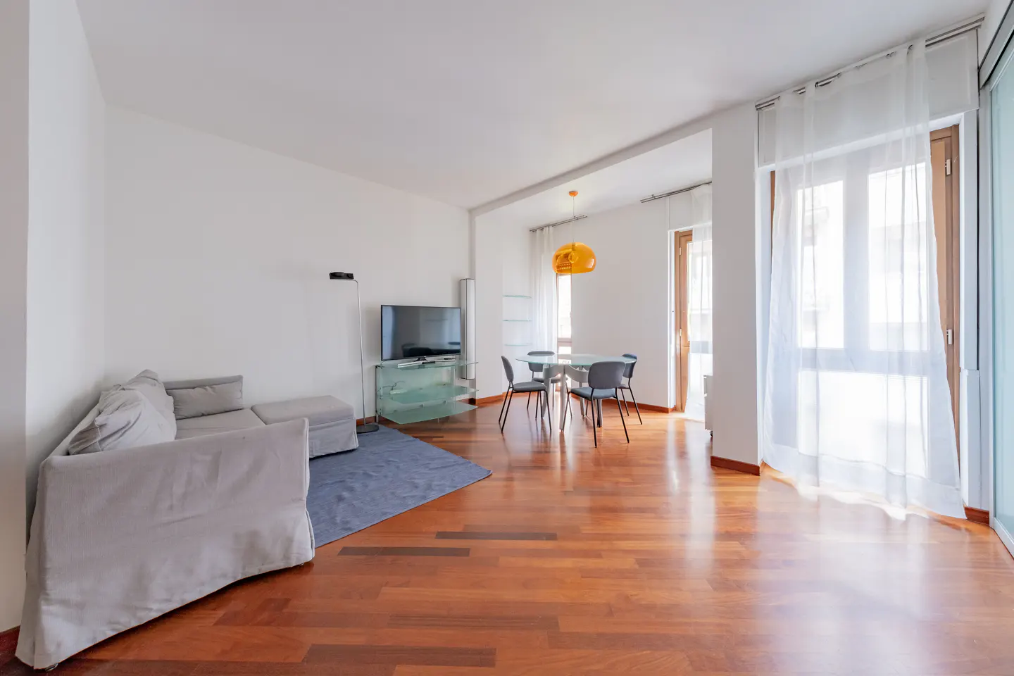 Bright living room with hardwood floors, white walls, and sheer curtains. A gray sofa sits near a TV, and a glass table with chairs is under an orange pendant light.