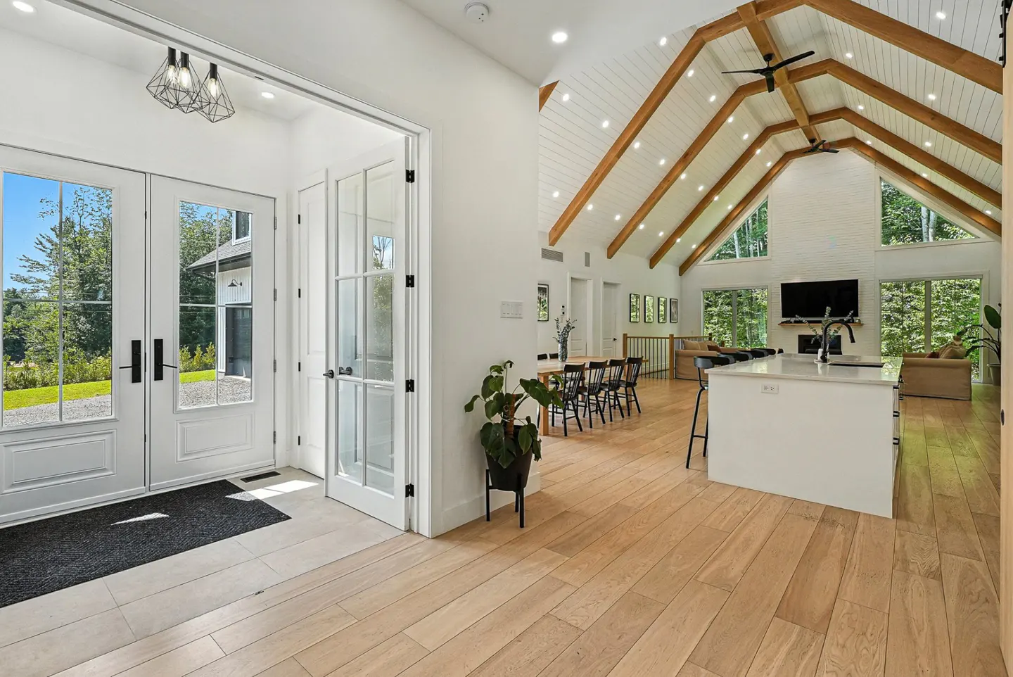Bright, open-concept home interior with white walls, wood floors, and vaulted ceiling with exposed beams. Double doors lead to the outside.
