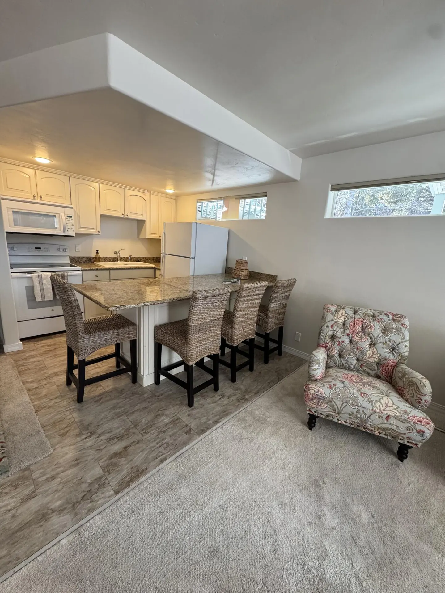 Open-concept kitchen and living area with white cabinets, island with wicker chairs, and floral armchair on gray carpet.