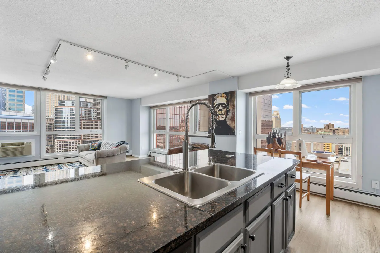 Open-concept condo kitchen with granite island, stainless steel sink, and city views from large windows. A dining table and living area are visible.