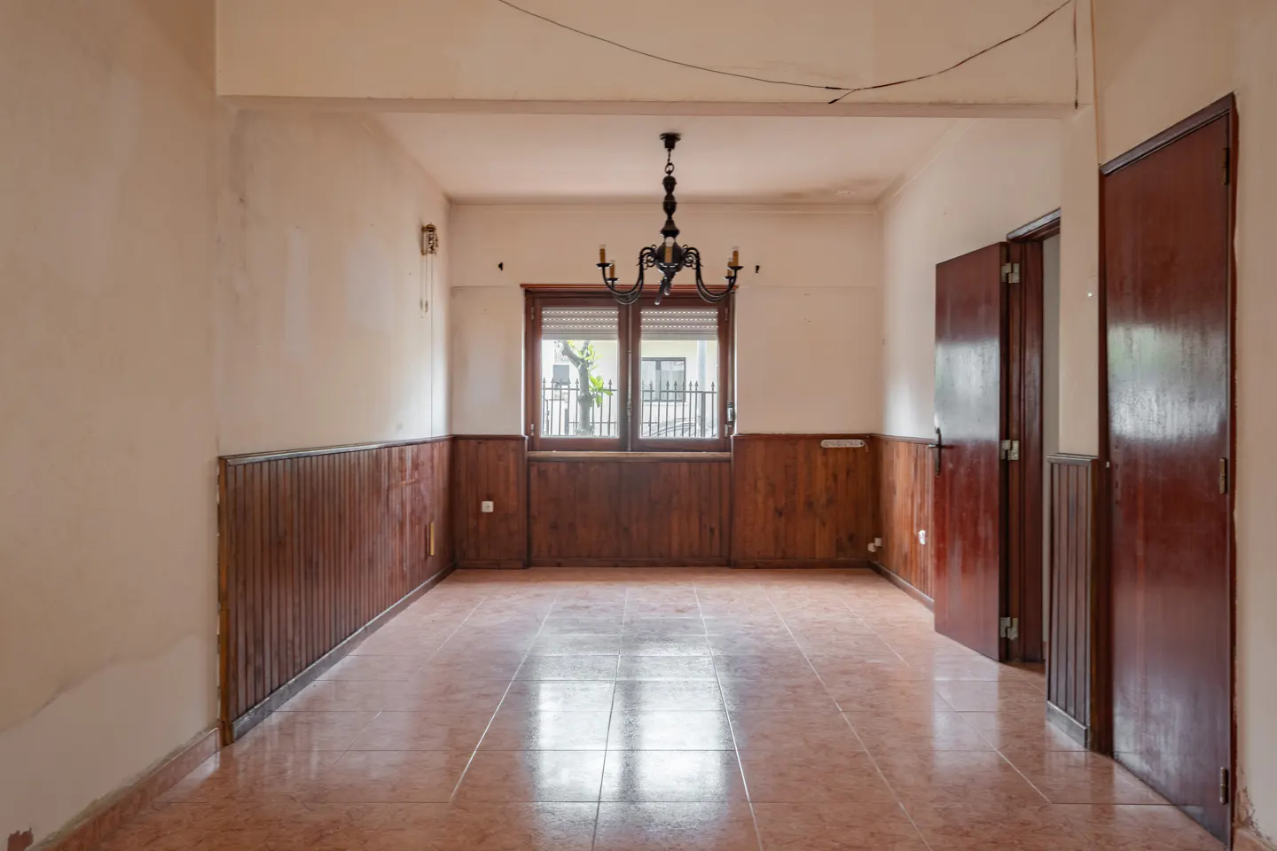 An empty room with wood paneling, tile floors, and a black chandelier. A window is centered on the back wall, and two doors are on the right.