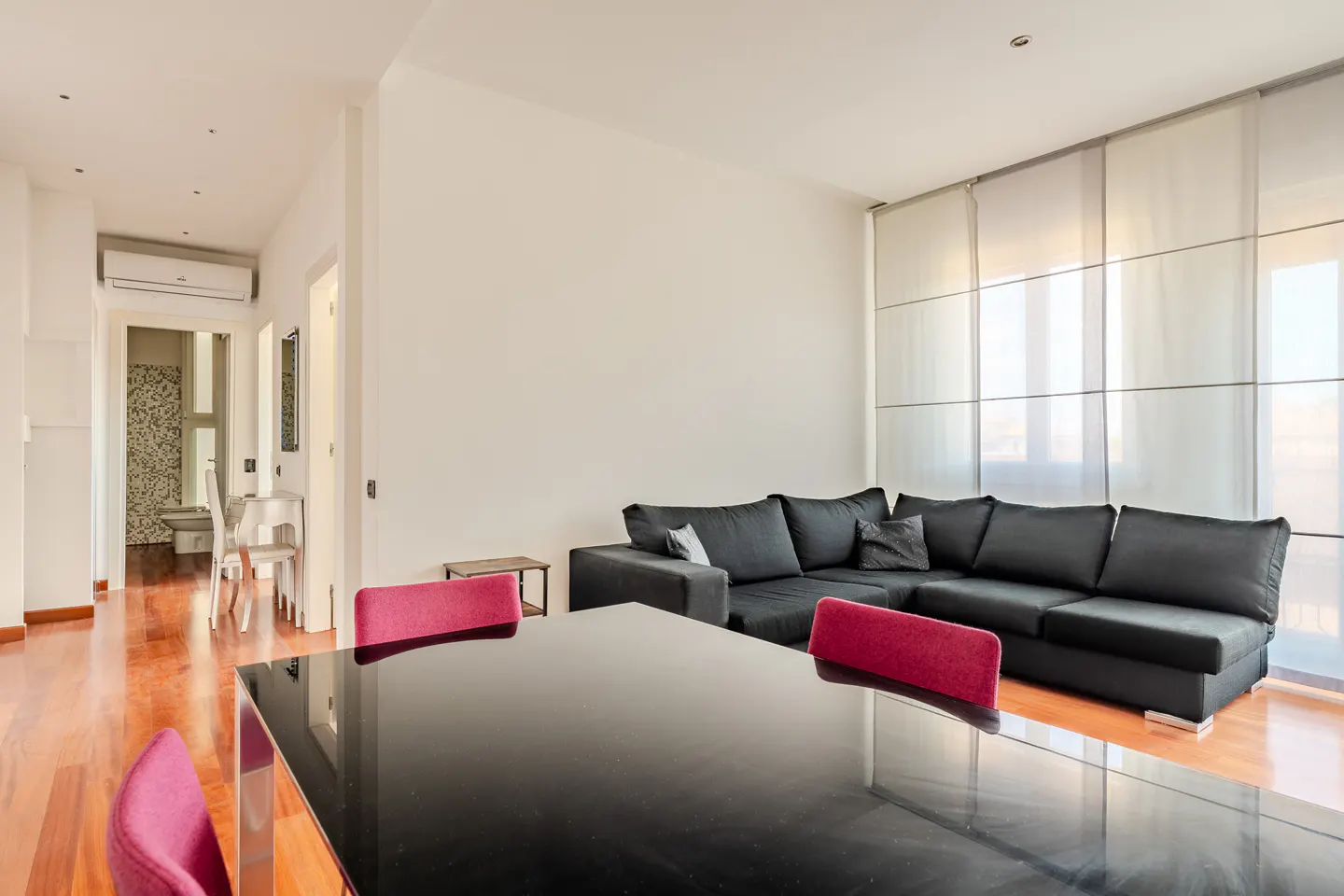 Bright living room with hardwood floors, a black sectional sofa, and a black dining table with red chairs. Large windows with white blinds.