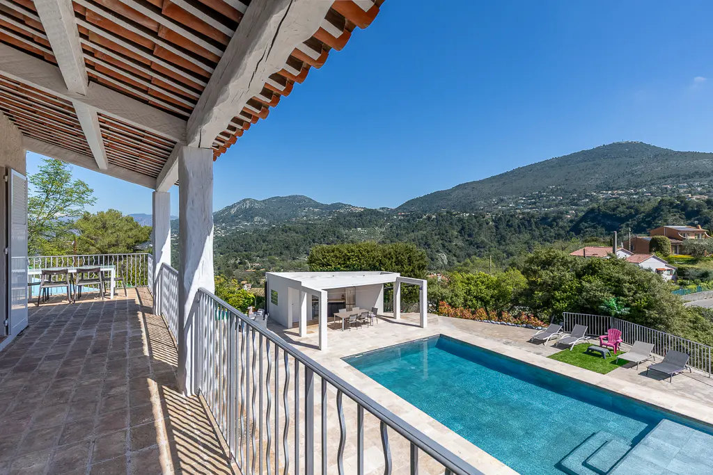 View from a villa balcony overlooking a pool, mountains, and blue sky. The balcony has a tiled floor and white railings.