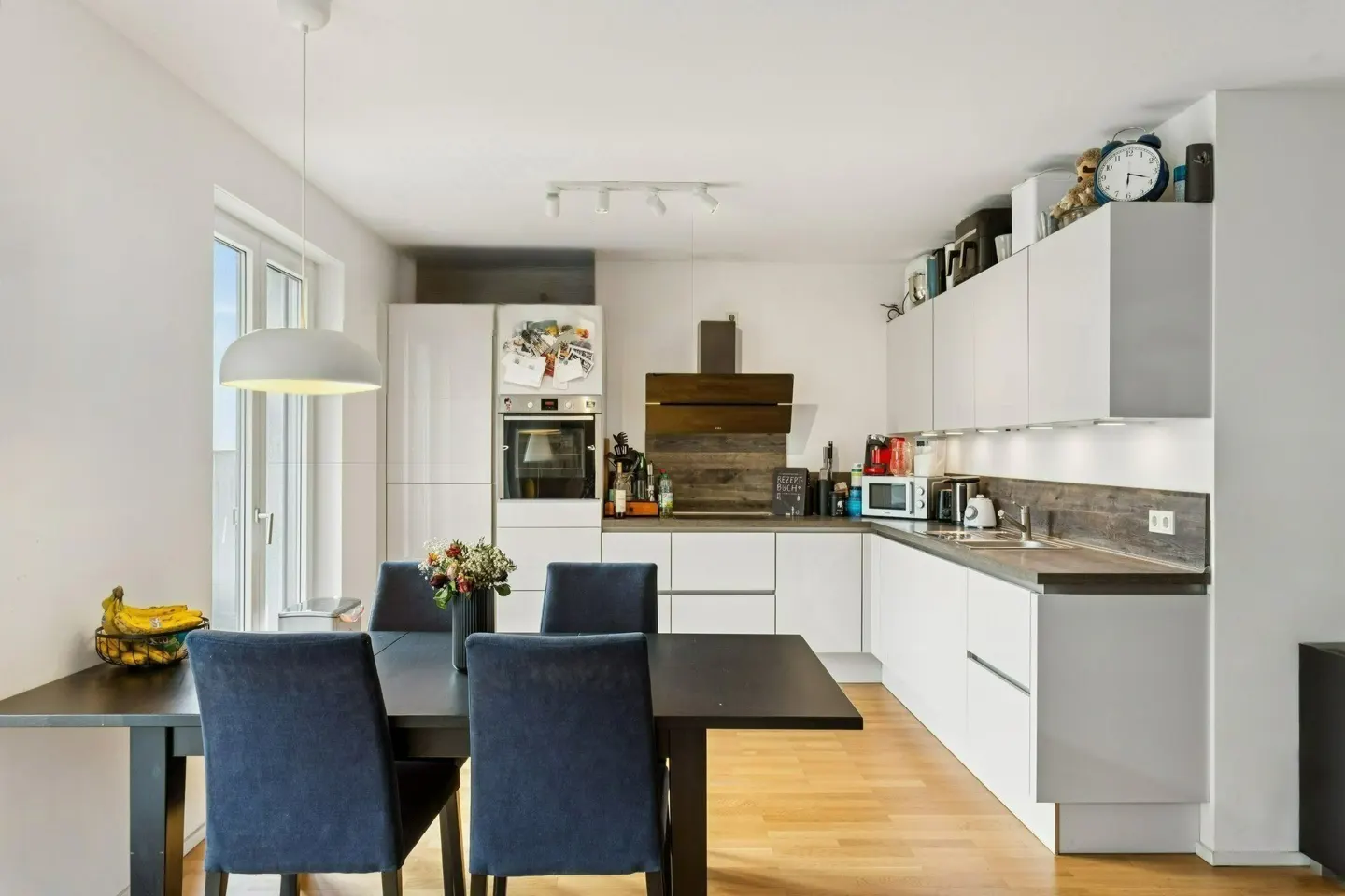 Modern kitchen with white cabinets, wood backsplash, and a dining area featuring a black table and four blue chairs.