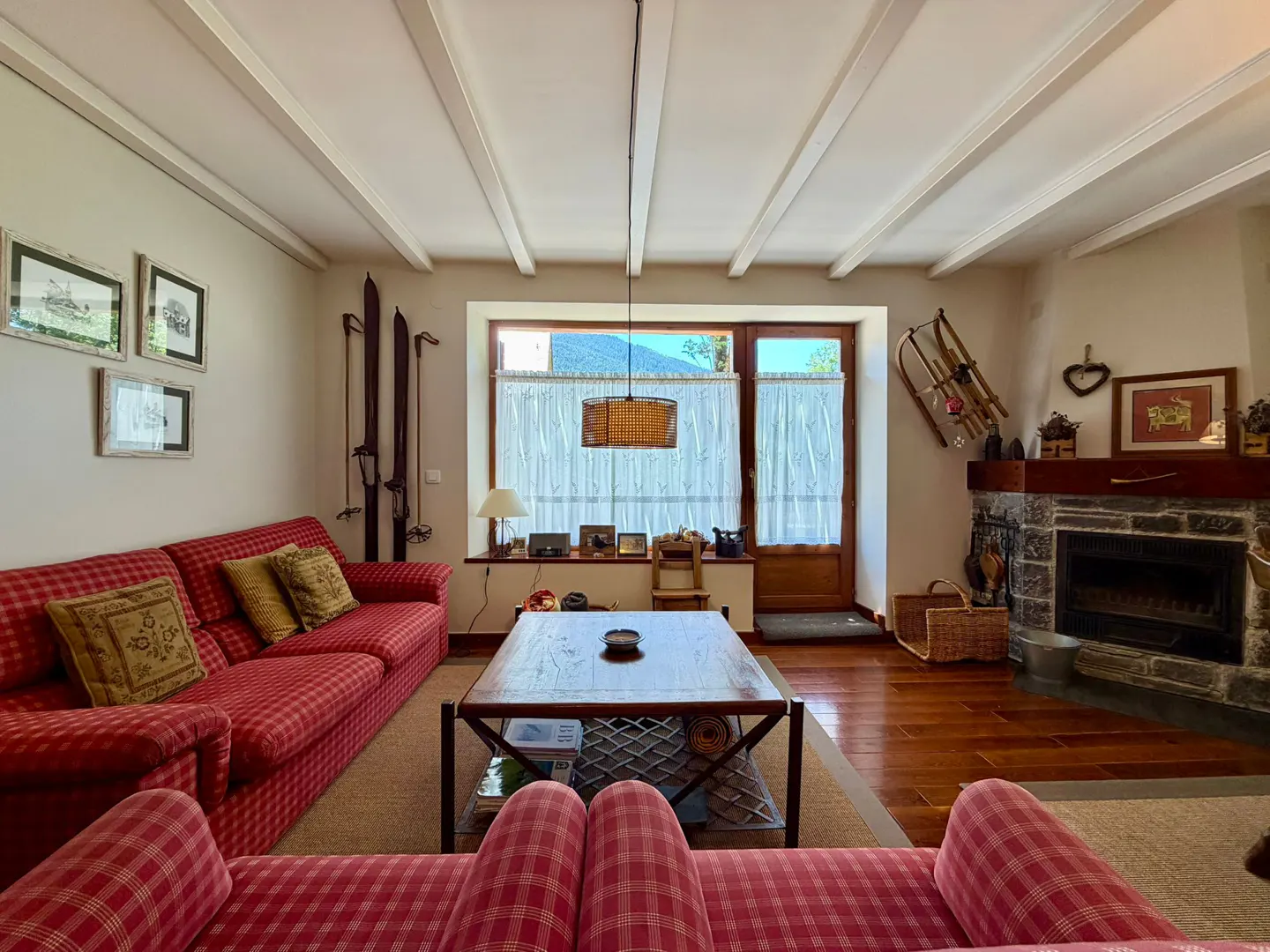 Living room with red plaid sofas, wood floors, and a stone fireplace. Skis and a sled decorate the walls. A window with white curtains is centered.