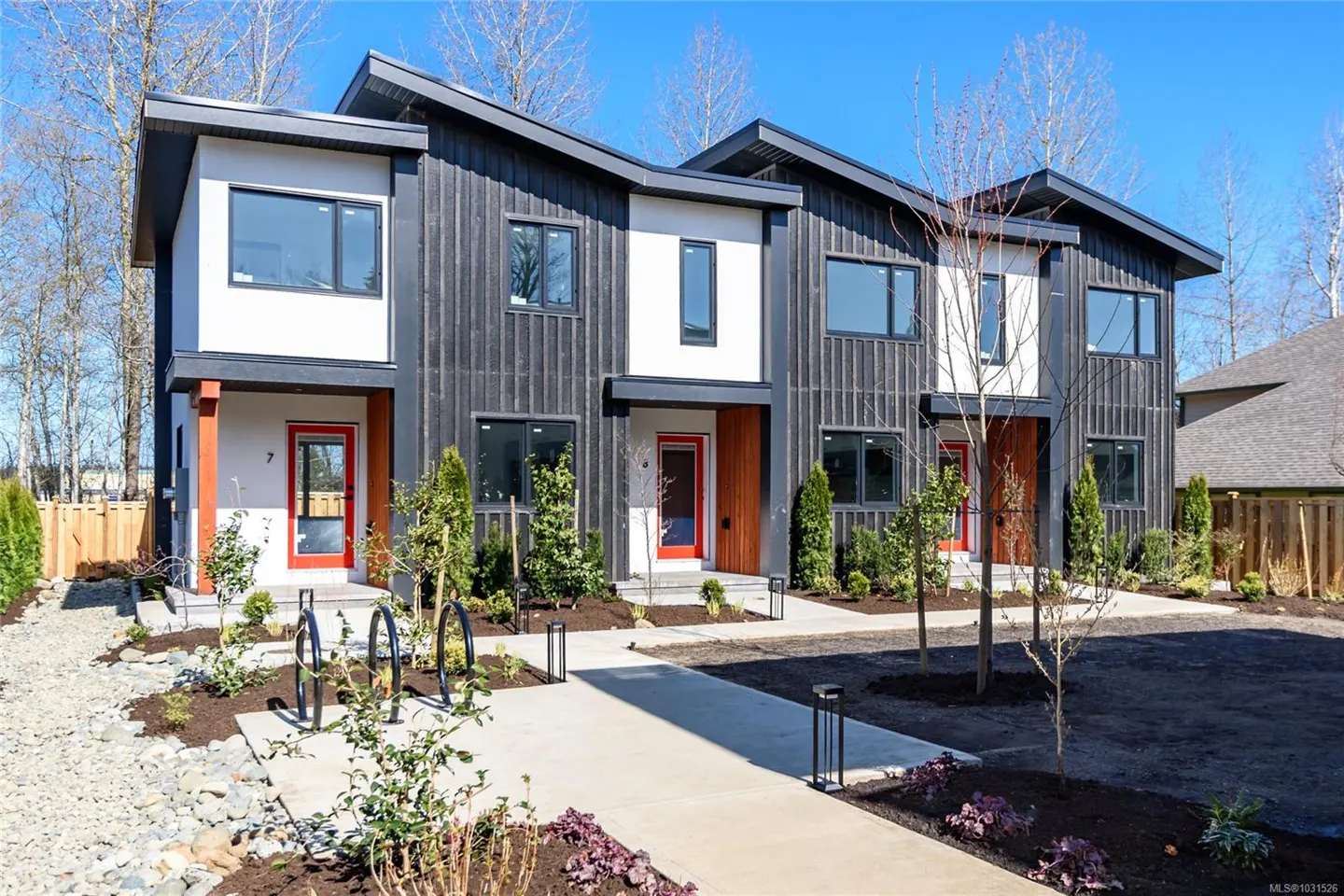 Modern townhouses with black siding, white accents, and red doors. A concrete walkway leads to the entrances, surrounded by landscaping.