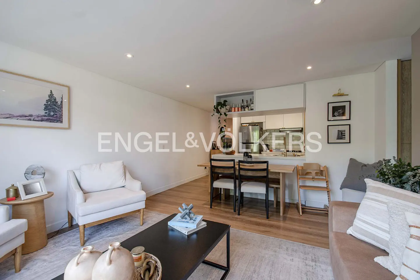 Bright living room with white walls, wood floors, and a black coffee table on a patterned rug. A dining table and chairs sit near a white kitchen.