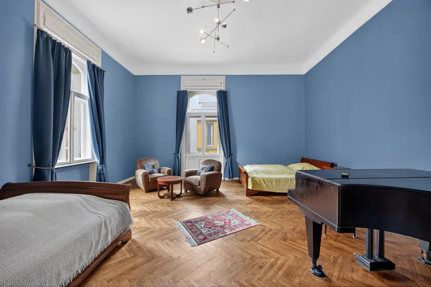 Bedroom with blue walls, herringbone wood floors, two beds, a piano, and a seating area with two armchairs.