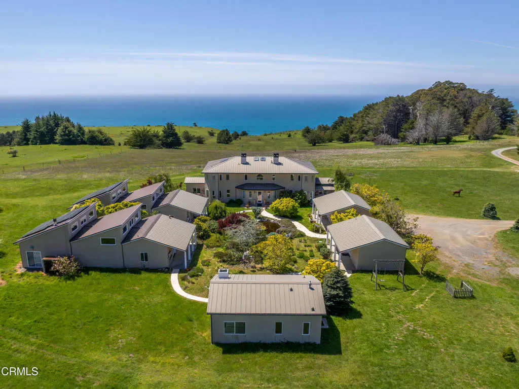 Aerial view of a large, light gray house with several smaller buildings surrounding it on a green lawn, with the ocean in the background.