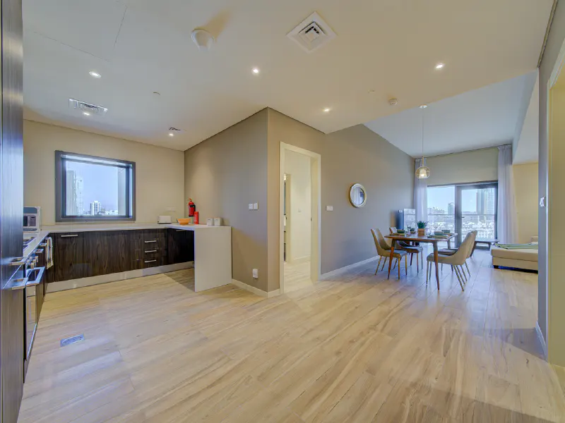Open-concept apartment with wood-look tile flooring, a kitchen with dark cabinets, and a dining area with a table and four chairs. A city view is visible through the window.