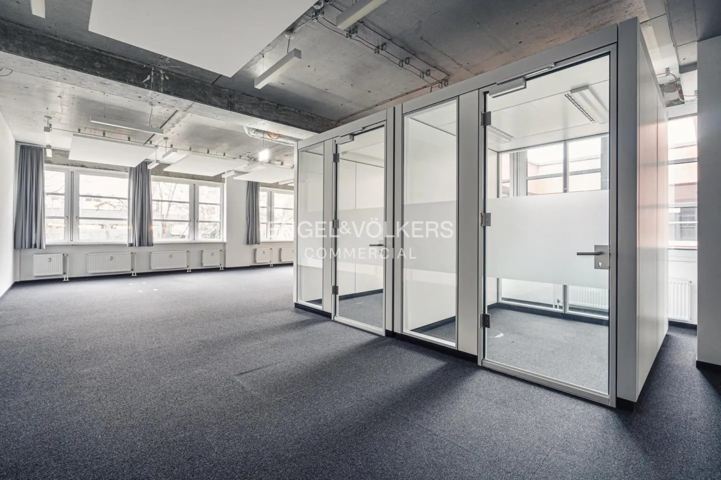 Empty office space with gray carpet, white walls, and glass-walled cubicles. Windows line the left wall with gray curtains. Concrete ceiling with lights.