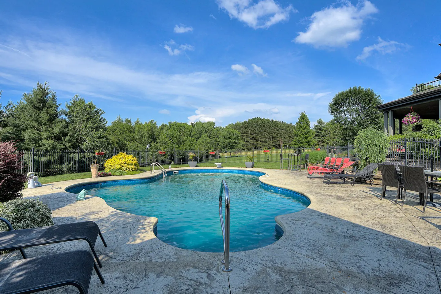 Outdoor pool with blue water, surrounded by a concrete patio, lounge chairs, and a green lawn with trees under a blue sky.