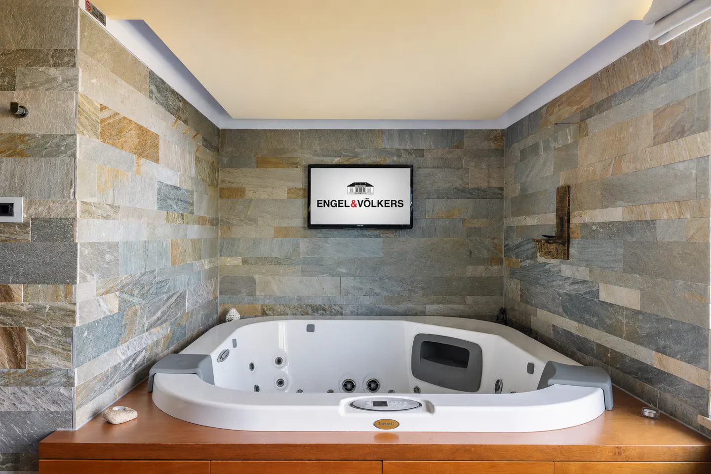 White hot tub set in a wooden base against a textured stone wall with a mounted television displaying a brand logo.