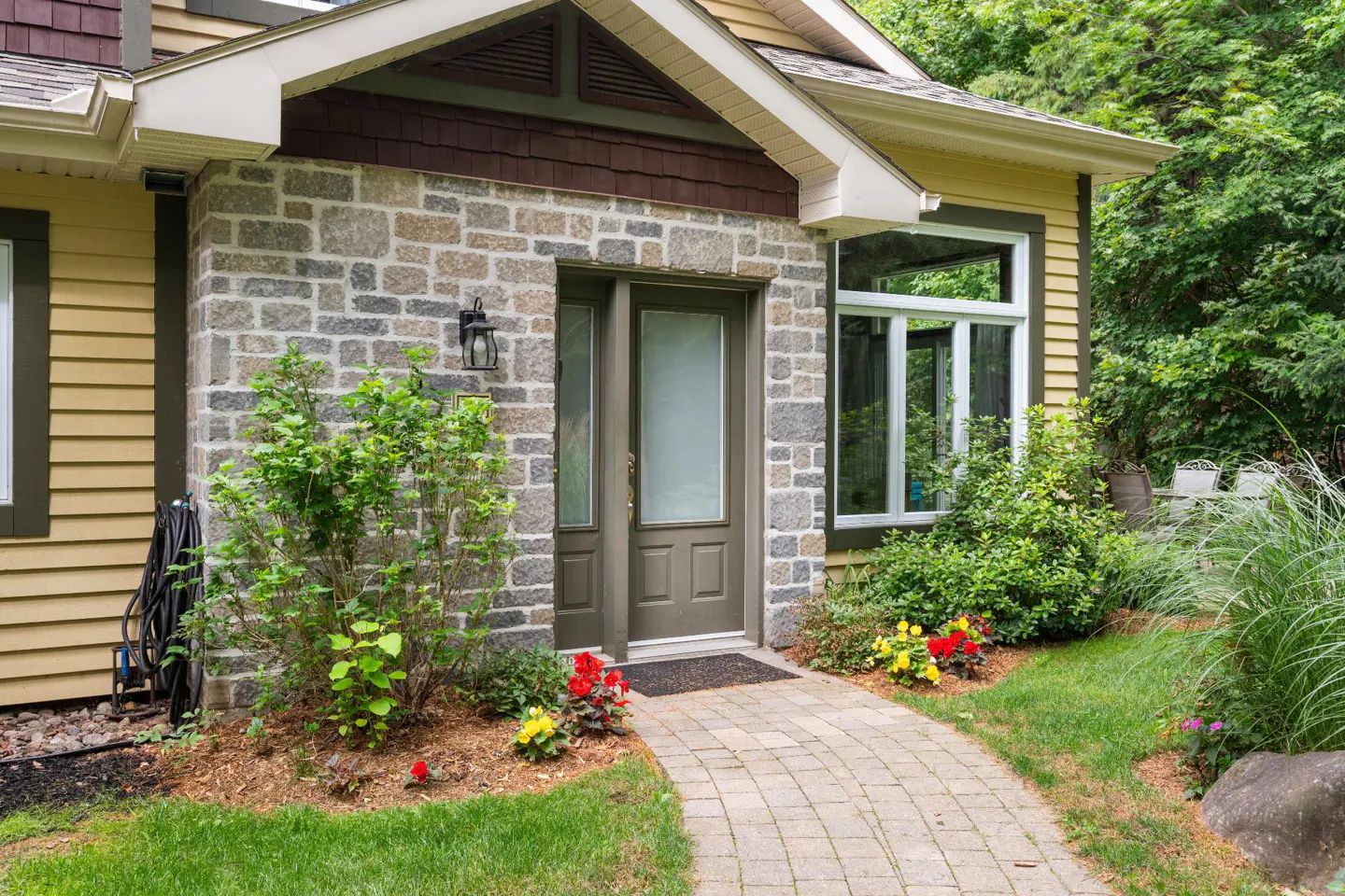 Exterior of a home with a stone facade, brown door, and a brick walkway leading to the entrance. Green foliage surrounds the path.