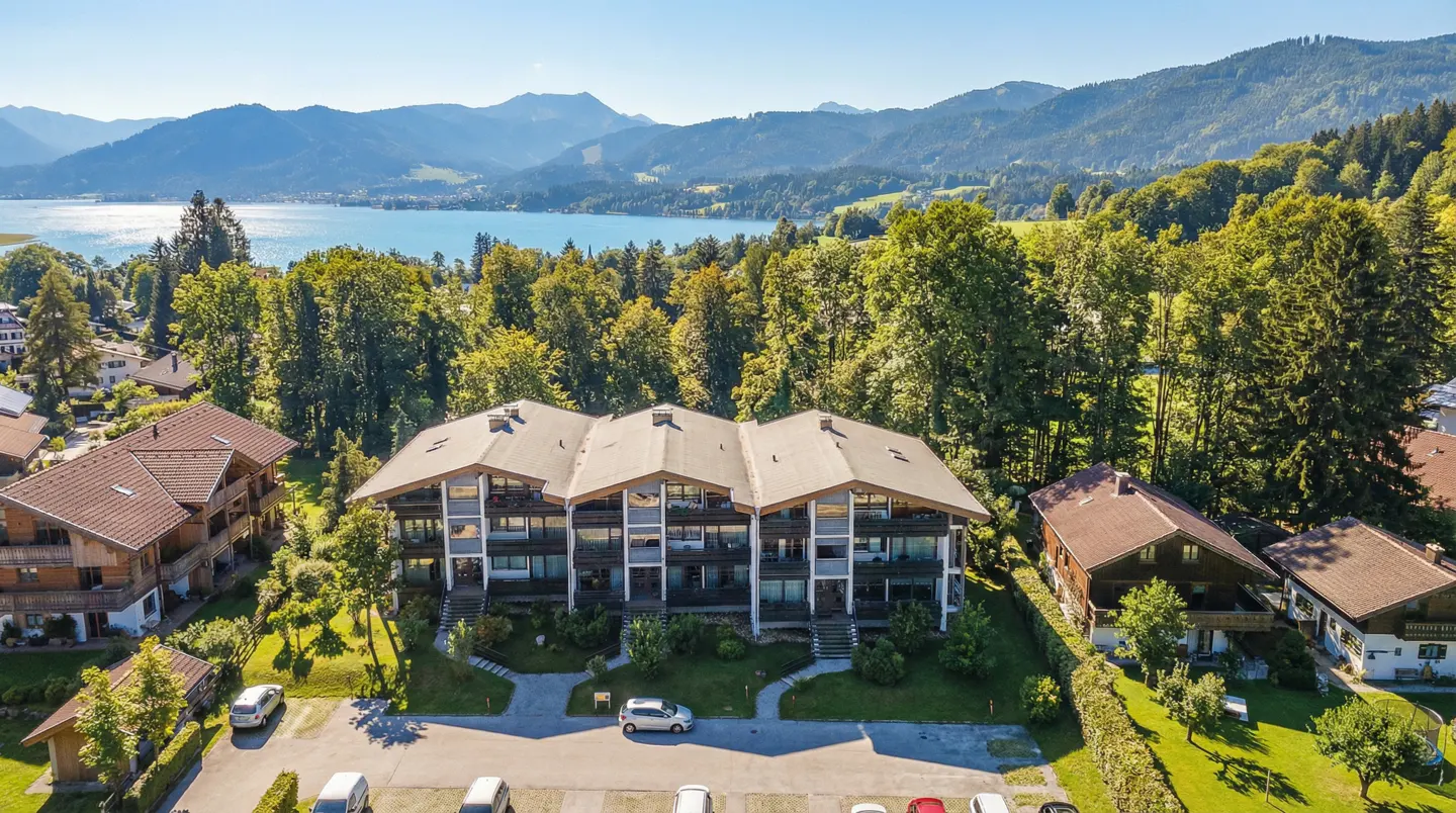 Aerial view of a three-story apartment building with a parking lot, surrounded by green trees, a lake, and mountains.
