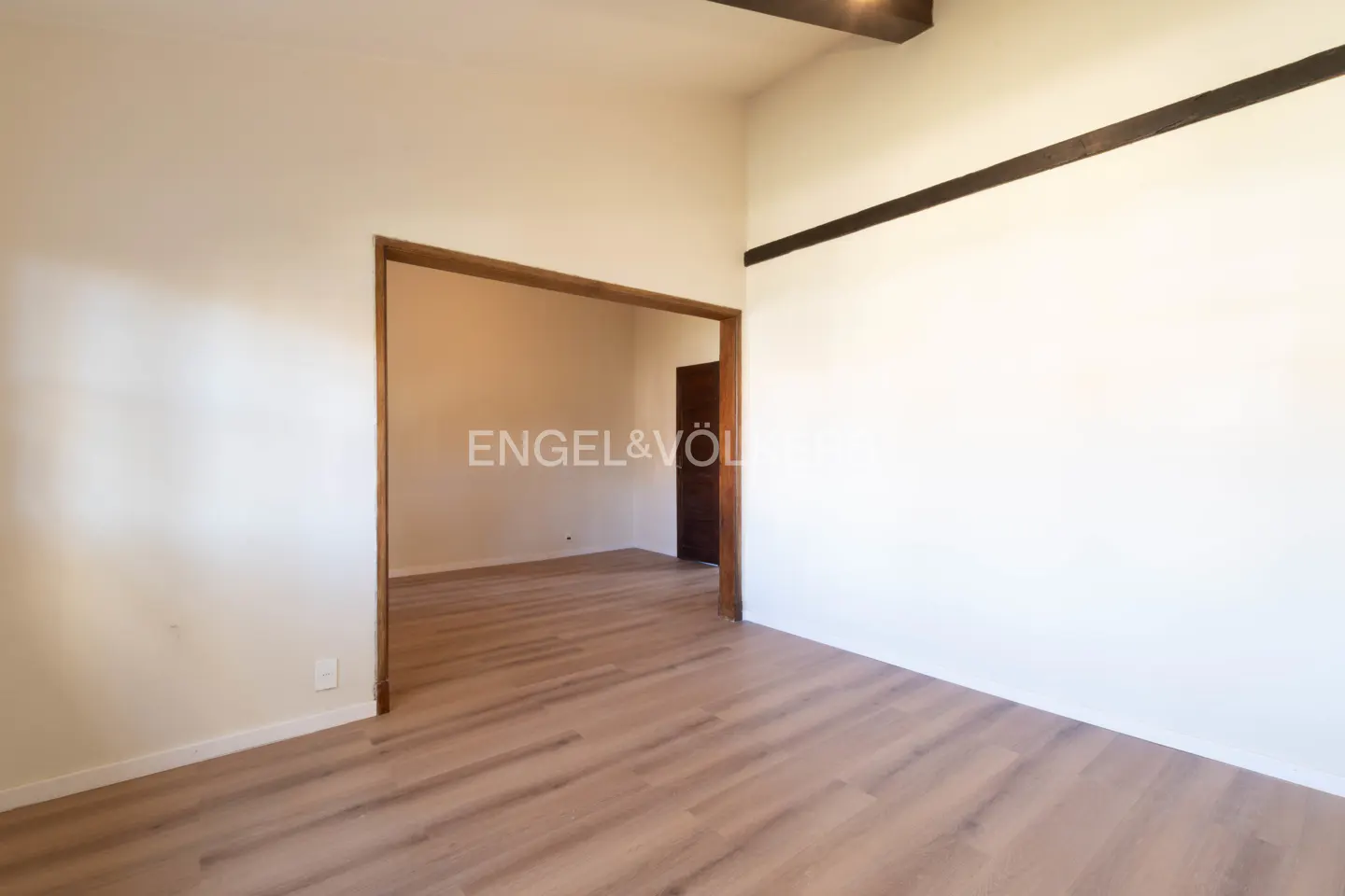 Empty room with light wood floors, cream walls, and a doorway to another room. A dark wood beam is visible on the ceiling.