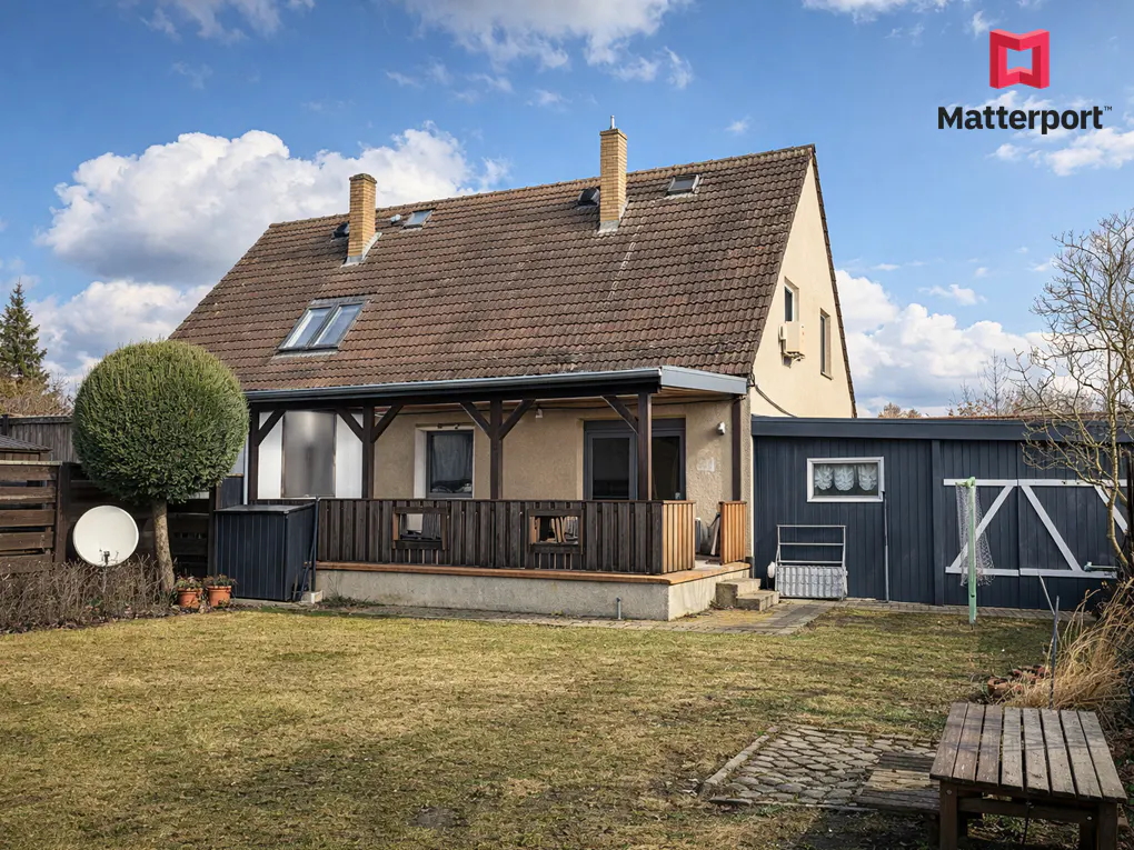 Backyard view of a tan house with a brown roof, a wooden deck, and a blue shed under a partly cloudy sky.