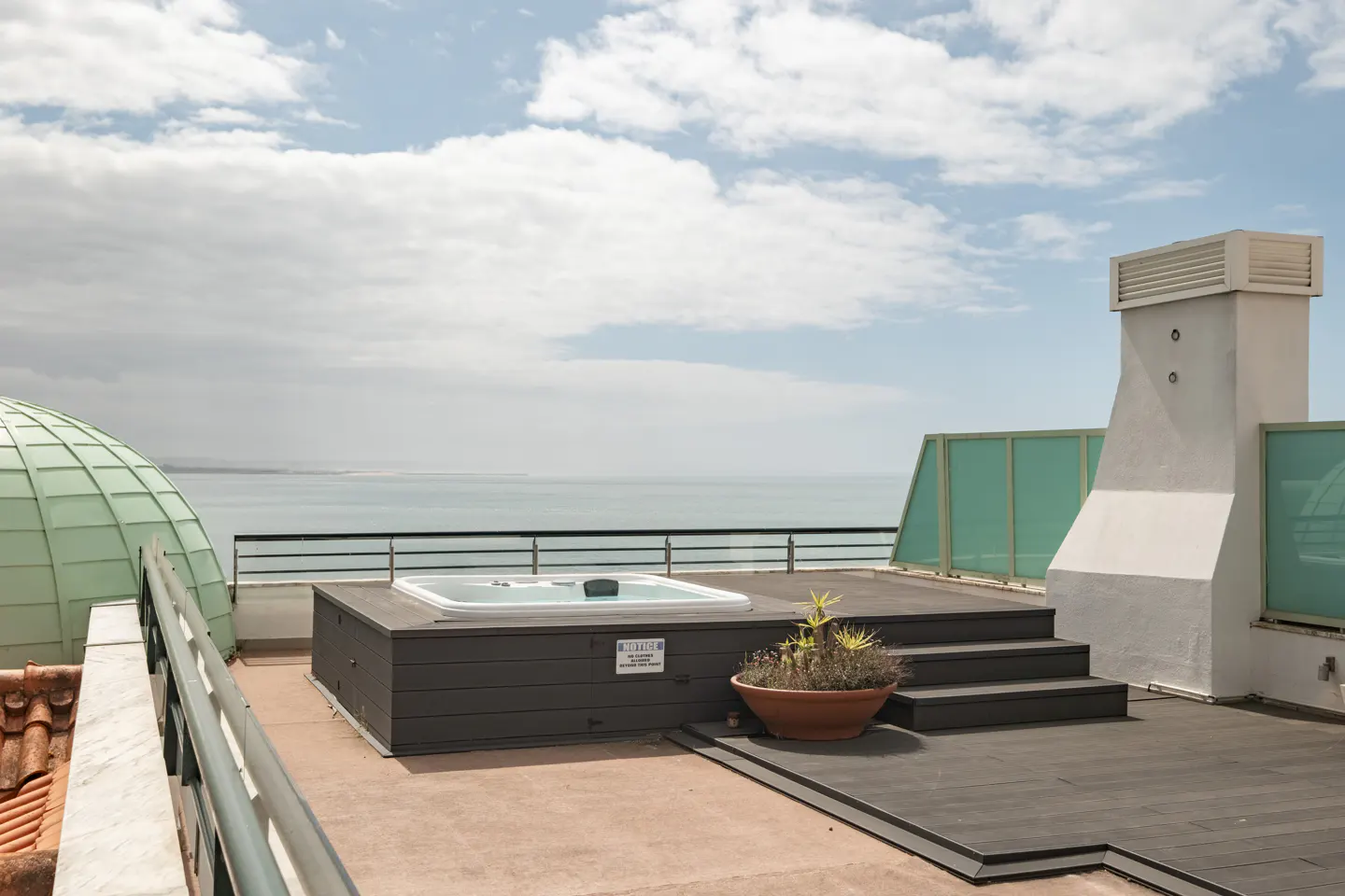 Rooftop view with a hot tub, ocean, and cloudy sky. A potted plant sits near the tub, with a white chimney and green dome in the background.