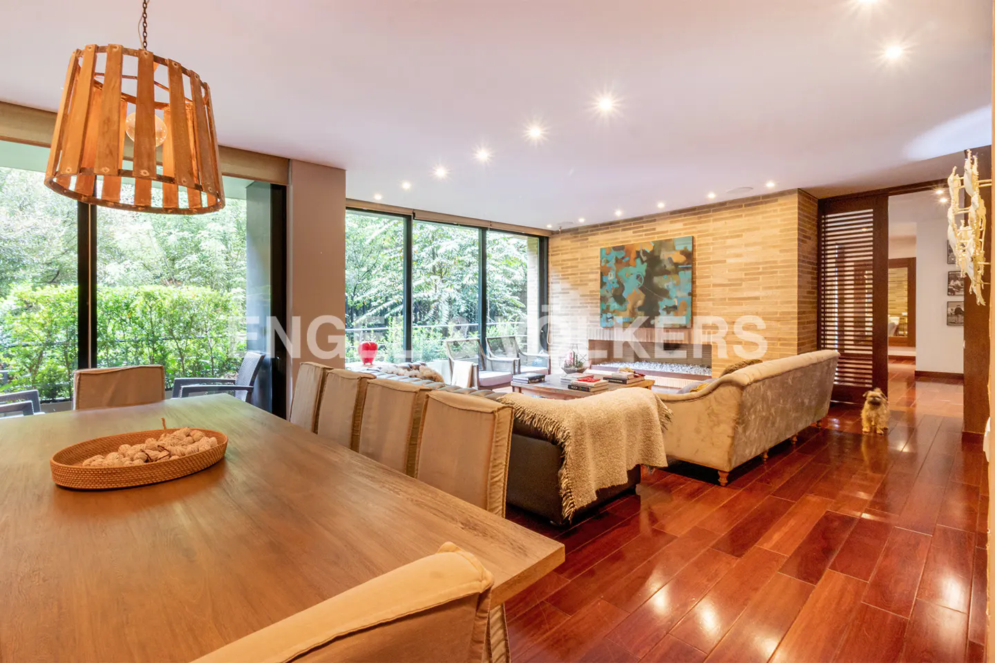 A spacious living room with a long wooden table, beige chairs, and a large window overlooking a green garden.
