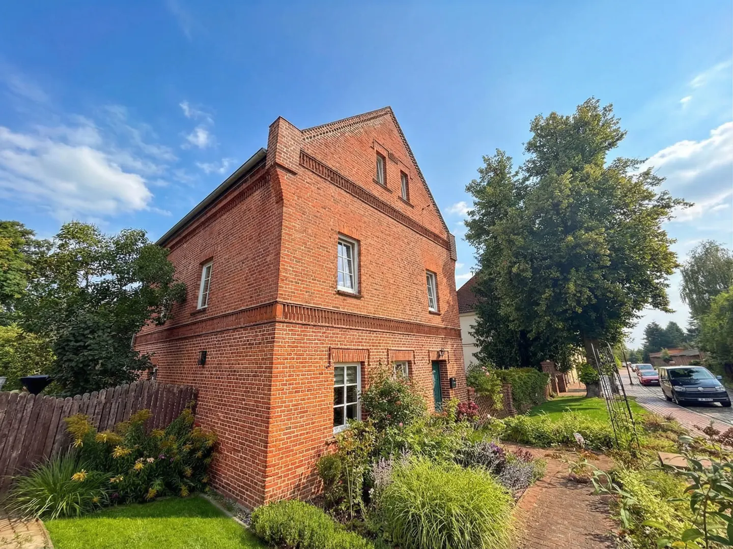 Exterior view of a two-story red brick house with white-framed windows, green door, and landscaped garden under a blue sky.