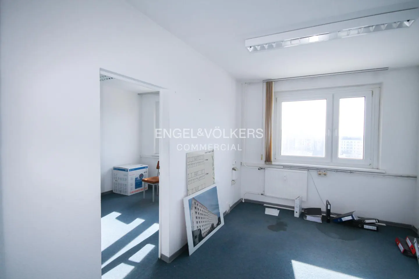 Bright, empty office space with white walls, blue carpet, and a large window. A doorway leads to another room with a box and chair. Engel & Völkers logo visible.