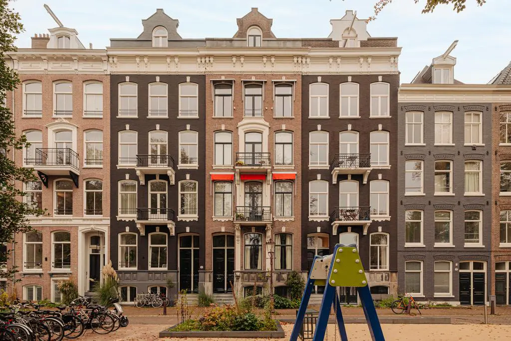 Row of tall, narrow brick buildings in Amsterdam, with white-framed windows and small balconies. A playground is in the foreground.