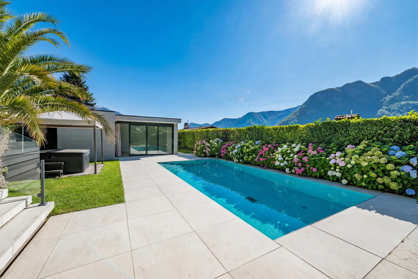 Outdoor pool with clear blue water, surrounded by white tiles, green hedge, colorful flowers, and mountains in the background.