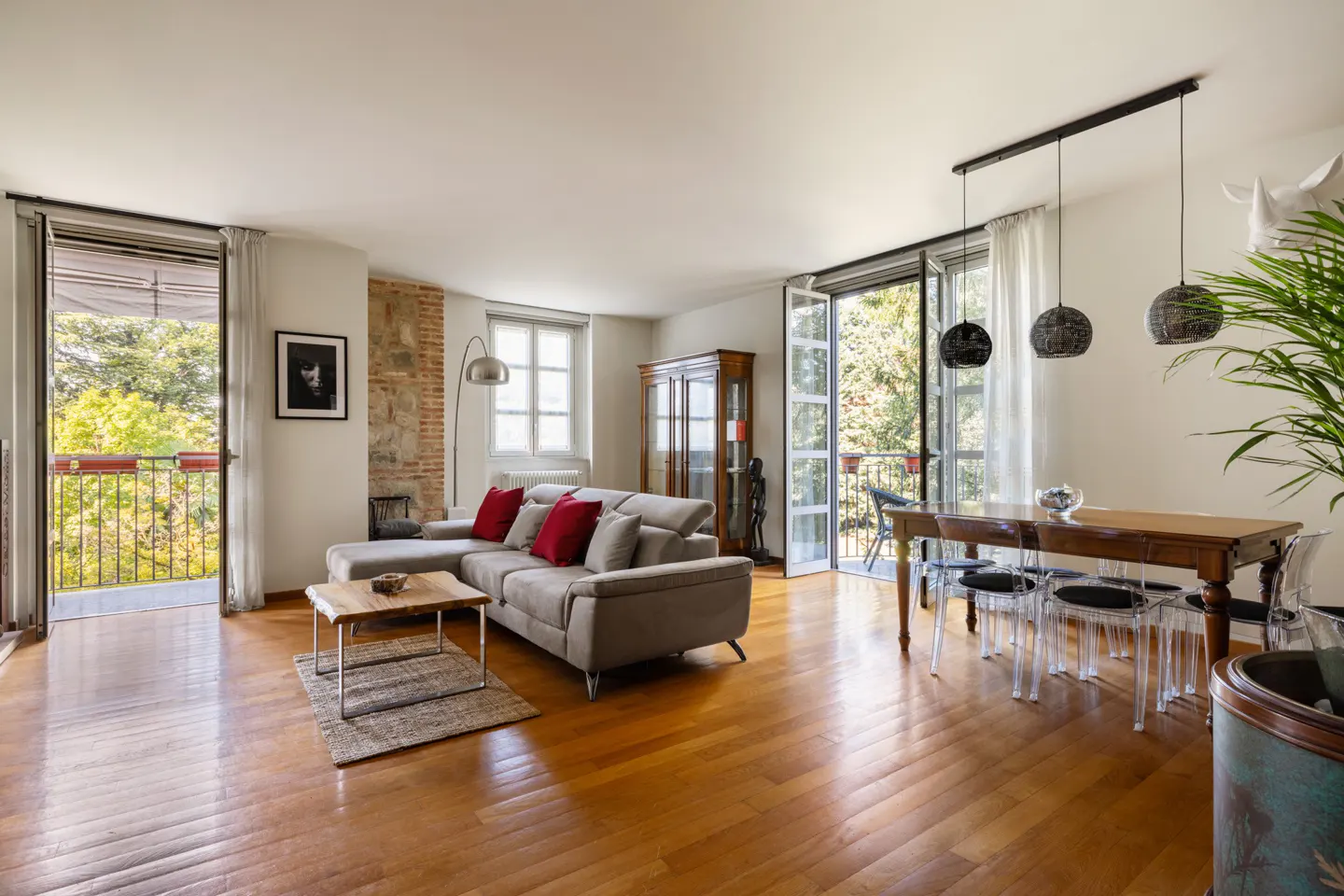 Bright living room with wood floors, a gray sectional sofa with red pillows, and a dining table with clear chairs. Balconies overlook green trees.