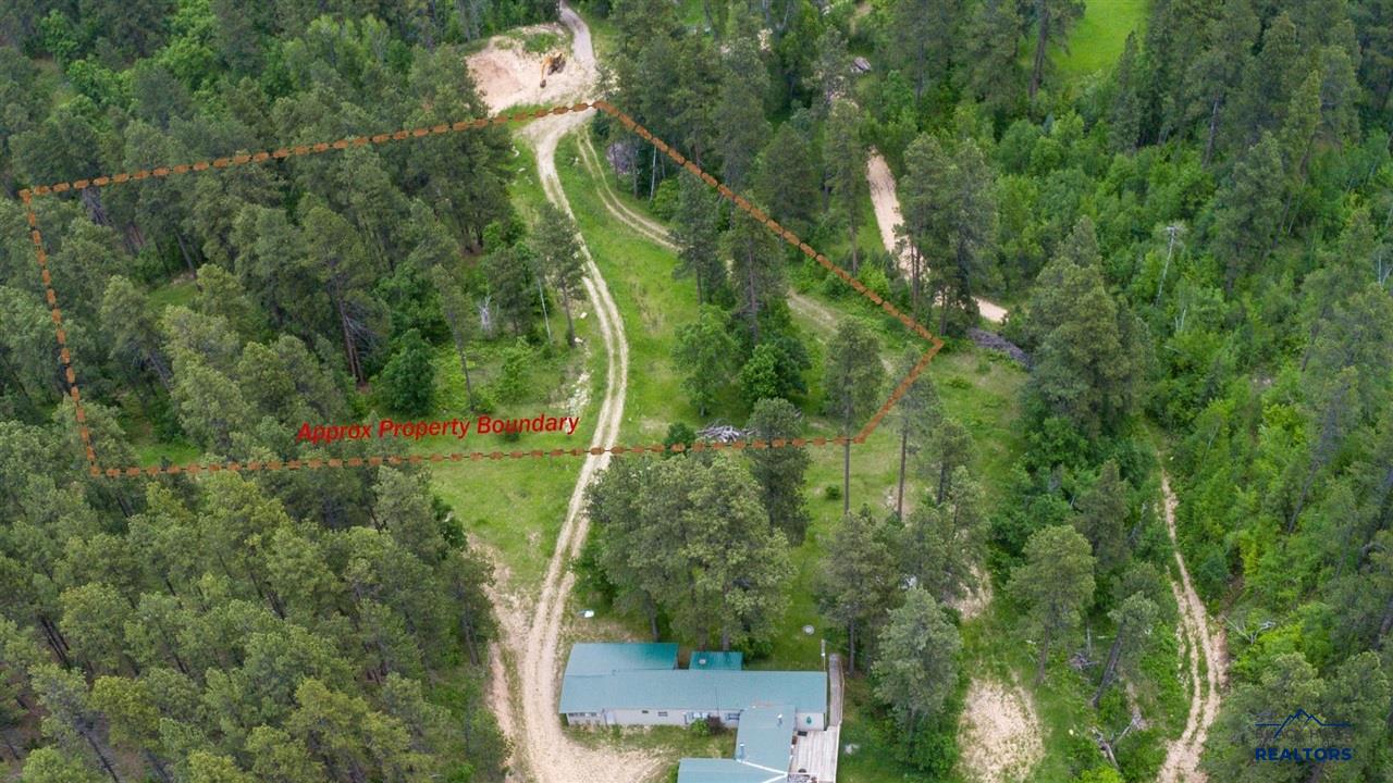 Aerial view of a wooded lot with a house, dirt driveway, and a dashed line marking the approximate property boundary. [AI-generated text]
