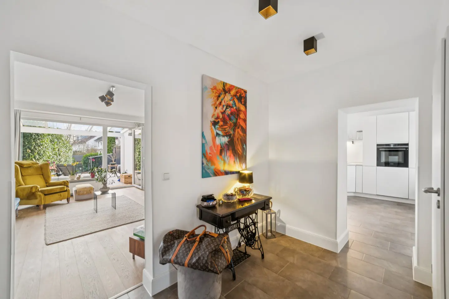 Hallway with white walls, tile floor, and doorways to a living room and kitchen. A lion painting hangs above a sewing machine table.