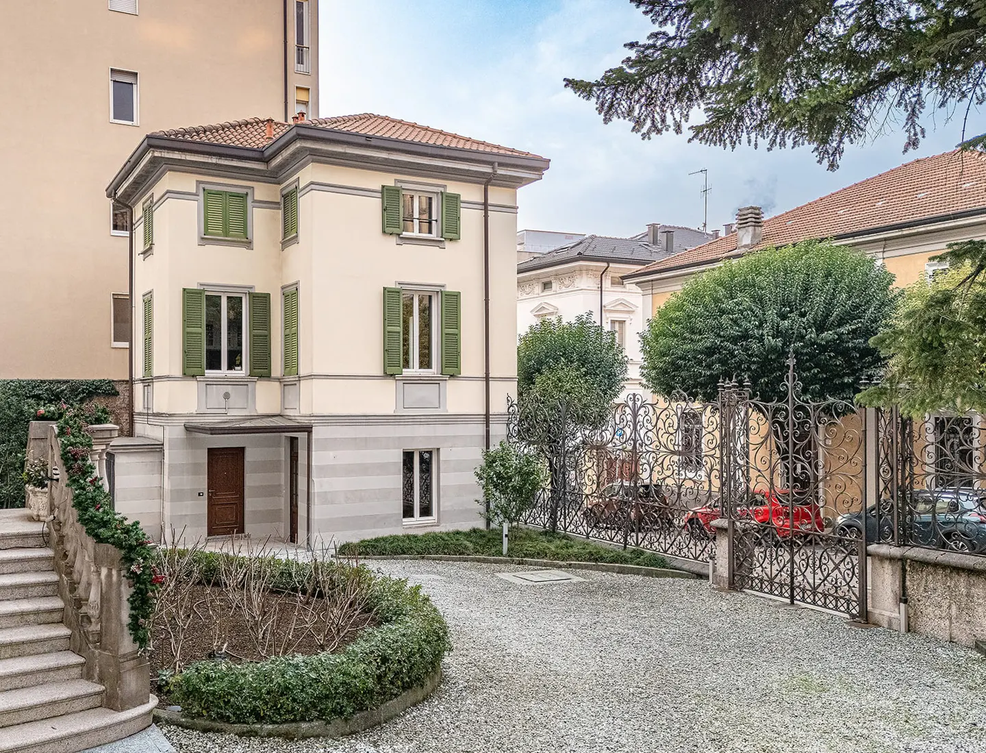 Two-story cream house with green shutters, a brown door, and a red tile roof. A gray gravel driveway leads to an ornate black metal gate.