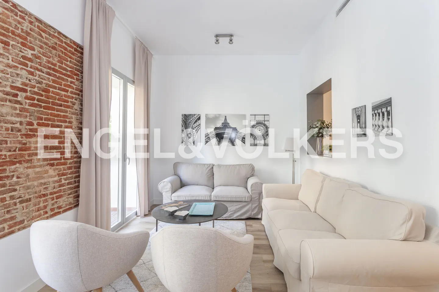Modern living room with a brick wall, beige sofas, two armchairs, a round coffee table, and Paris-themed wall art.