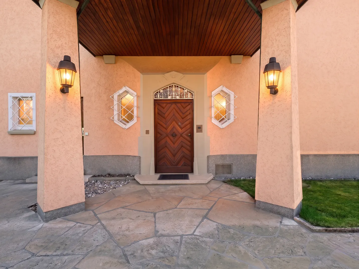 Front entrance of a peach-colored house with a wooden door, stone walkway, and lit lanterns.