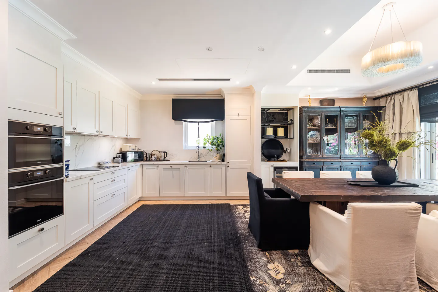 Bright kitchen with white cabinets, marble countertops, and black appliances. A dark wood table with white and black chairs sits on a floral rug.