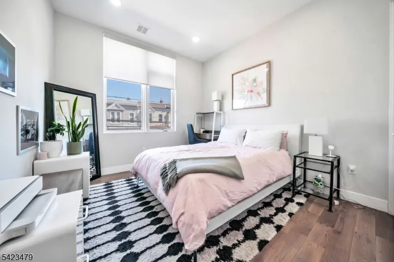 Bedroom with a pink bedspread, white walls, a black and white rug, and a window with a view of houses.