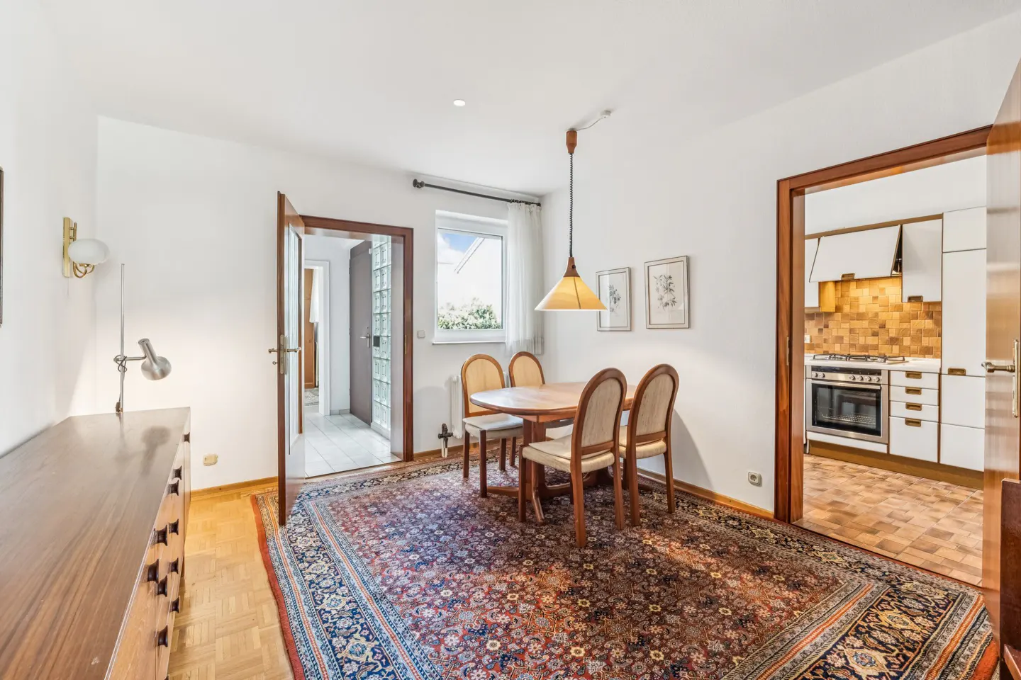Dining room with wooden table, four chairs, patterned rug, and pendant light, opening into a kitchen and hallway.