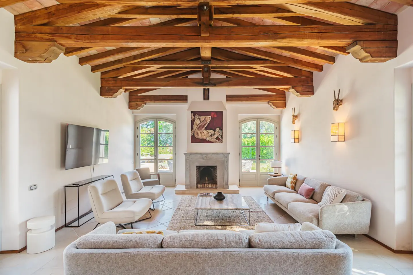Bright living room with exposed wood beams, white walls, and tile floors. Sofas and chairs surround a coffee table and fireplace.