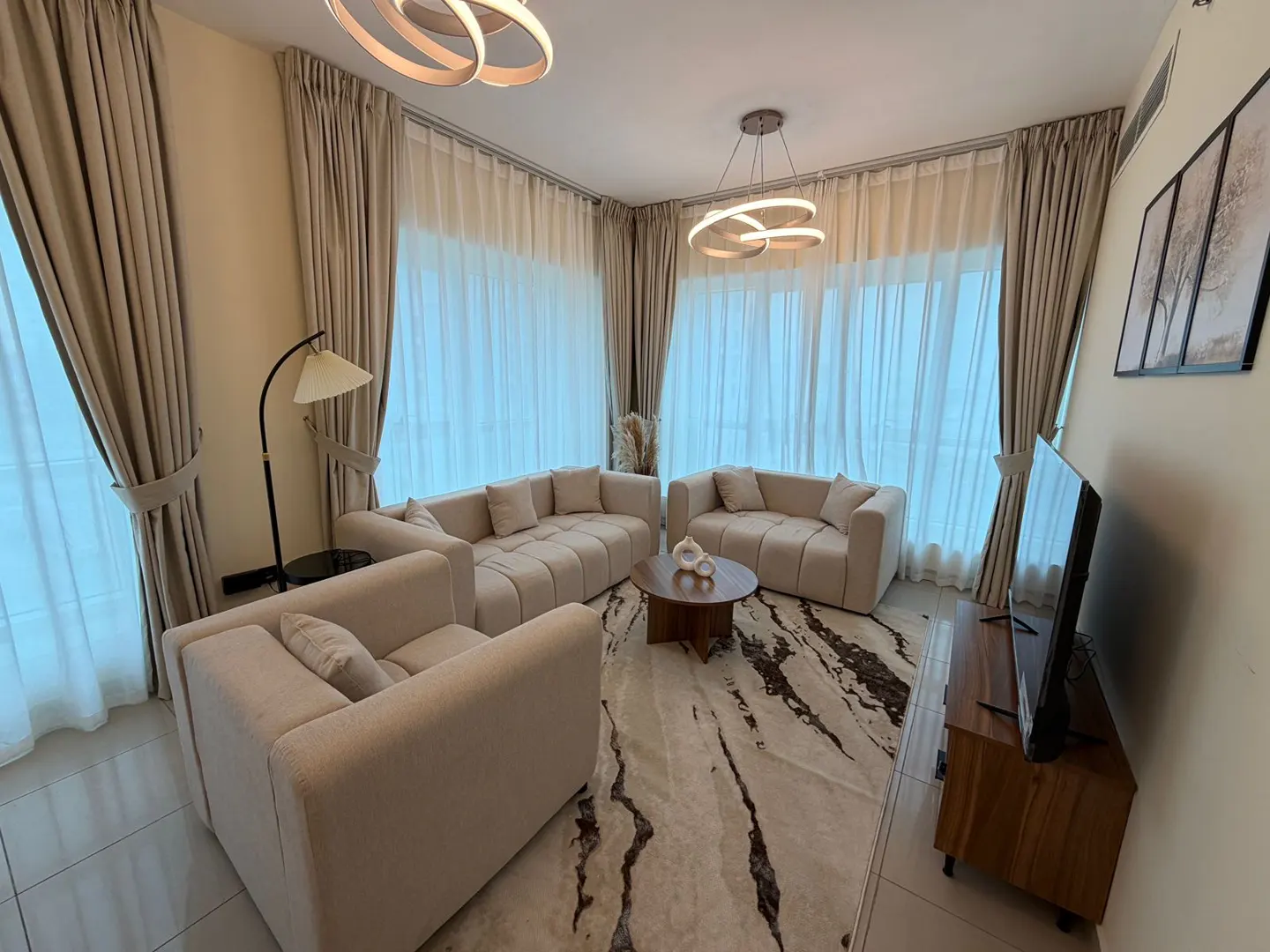 Living room with beige sofas, a round wood table, and a patterned rug. Light streams through sheer curtains. Modern lighting fixtures.