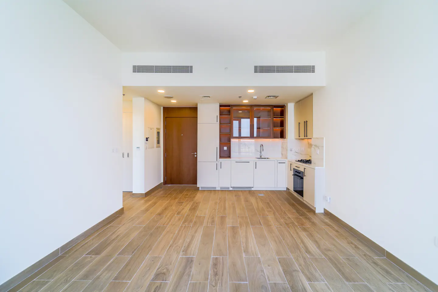 Bright, modern apartment interior with white walls, wood-look tile flooring, and a compact kitchen with white cabinets and stainless steel appliances.