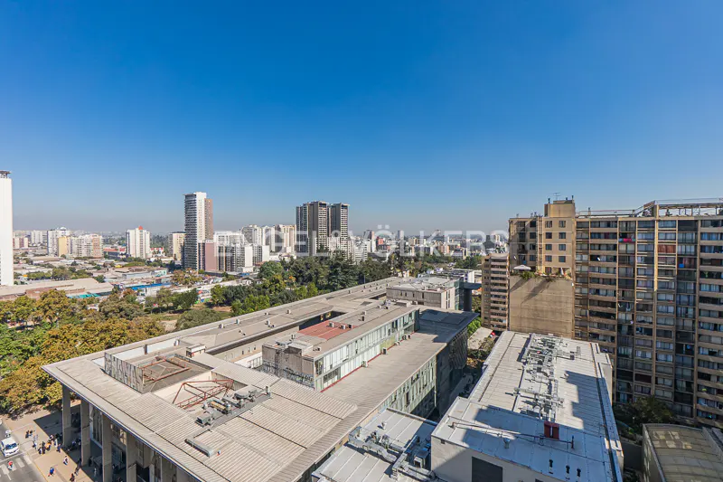 Cityscape view under a clear blue sky, featuring tall buildings, trees, and rooftops.