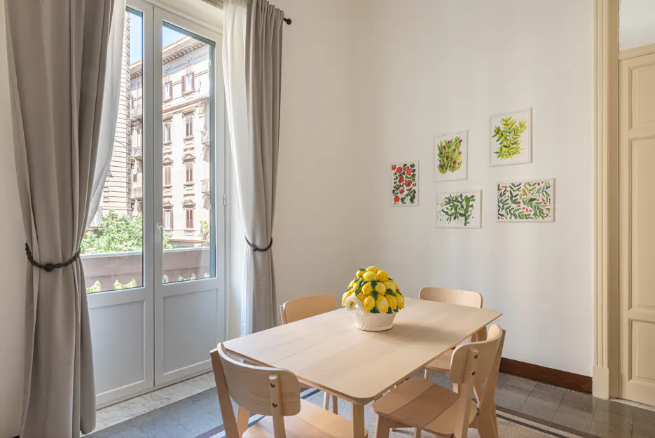 Bright dining room with a light wood table, chairs, and a vase of lemons. Botanical prints hang on the white wall. Balcony doors with gray curtains.